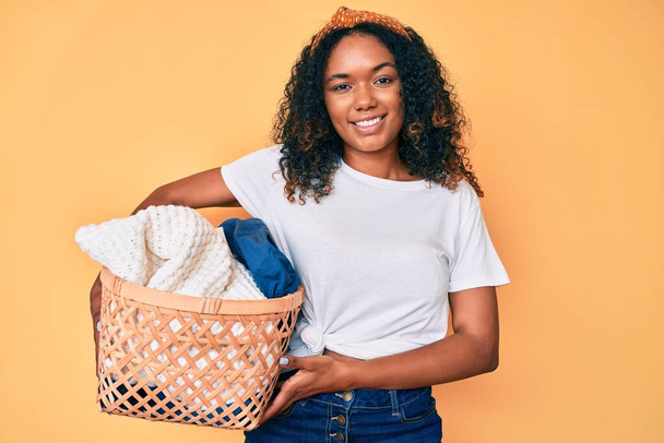 Joven mujer afroamericana sosteniendo cesta de ropa que se ve positiva y feliz de pie y sonriendo con una sonrisa confiada mostrando los dientes  - Foto, Imagen