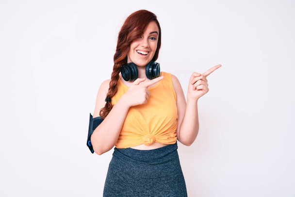 Young beautiful woman wearing gym clothes and using headphones smiling and looking at the camera pointing with two hands and fingers to the side.  - Photo, Image