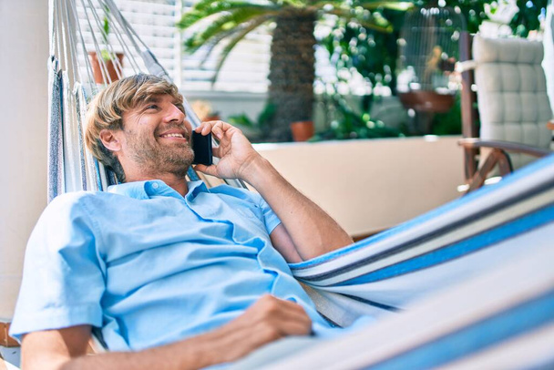 Średniego wieku przystojny mężczyzna na tarasie swojego domu relaksujący się leżąc na hamaku rozmawiając przez telefon - Zdjęcie, obraz