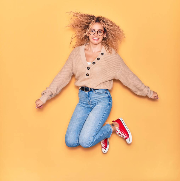 Young beautiful curly girl wearing casual clothes and glasses smiling happy. Jumping with smile on face and arms opened over isolated yellow background. - Foto, afbeelding