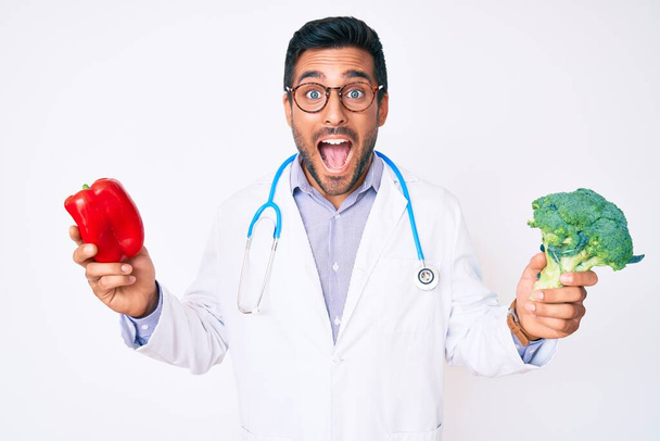 Young hispanic man wearing doctor stethoscope holding red pepper and broccoli celebrating crazy and amazed for success with open eyes screaming excited.  - Photo, Image