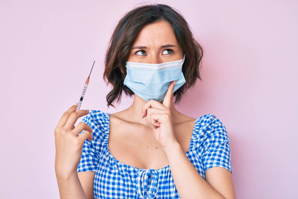 Молодая красивая женщина в медицинской маске держит шприц серьезное лицо думая о вопросе с рукой на подбородке, вдумчивый о запутанной идее  - Фото, изображение