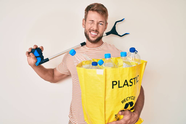 Fiatal kaukázusi férfi gazdaság újrahasznosító zsák műanyag palackok és hulladékszedő kacsintás a kamerába szexi kifejezés, vidám és boldog arc.  - Fotó, kép