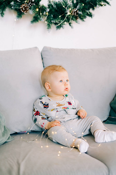 primeiro Natal da criança.um menino bonito está sentado em um traje festivo em um sofá cinza, contra uma parede branca com uma grinalda natural de abeto.. - Foto, Imagem