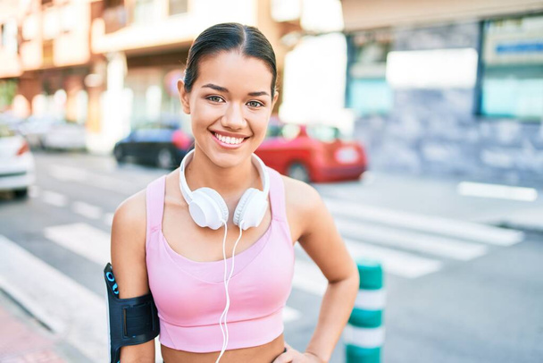 Junge schöne hispanische Sportlerin im Läufer-Outfit und Kopfhörer lächelt glücklich in die Stadt - Foto, Bild
