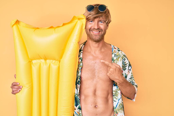 Giovane uomo biondo che indossa lo stile estivo e che tiene il galleggiante materasso sorridente felice indicando con mano e dito  - Foto, immagini