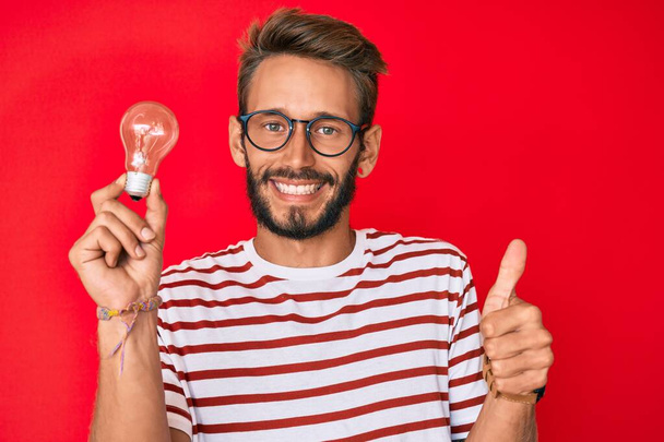 Handsome caucasian man with beard holding lightbulb for inspiration and idea smiling happy and positive, thumb up doing excellent and approval sign  - Photo, Image