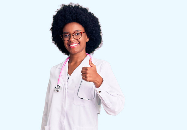 Młoda Afroamerykanka w płaszczu lekarskim i stetoskopie wykonująca gesty ręką w górę. zatwierdzanie ekspresji patrząc w aparat pokazujący sukces.  - Zdjęcie, obraz
