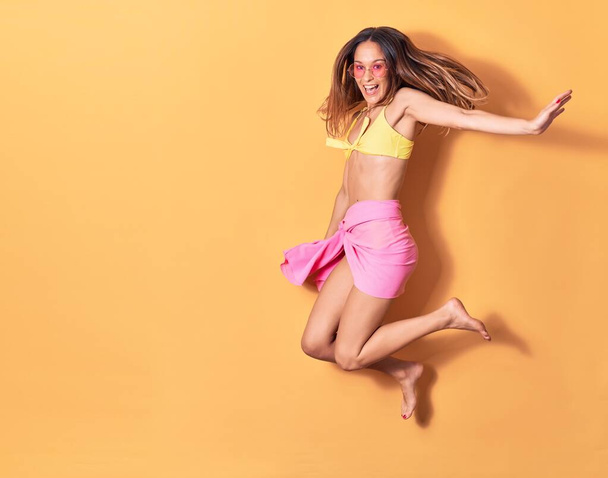 Young beautiful girl wearing bikini and sunglasses smiling happy. Jumping with smile on face over isolated yellow background - 写真・画像