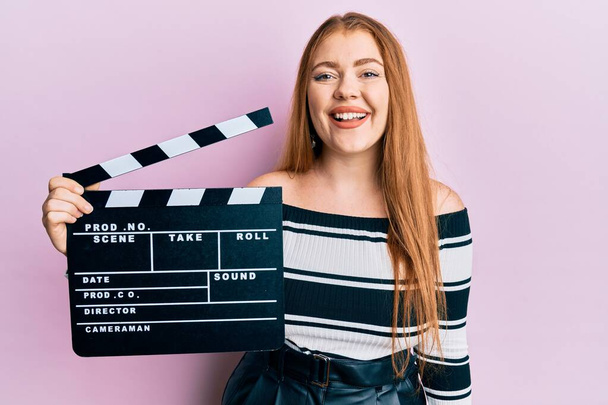 Jonge mooie roodharige vrouw met video film klapbord kijken positief en gelukkig staan en glimlachen met een zelfverzekerde glimlach tonen tanden  - Foto, afbeelding
