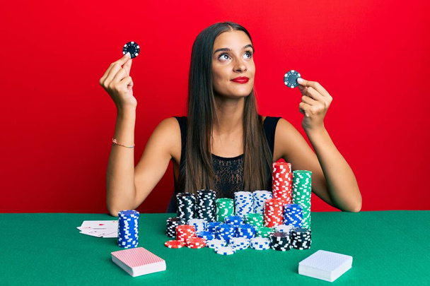 Jeune femme hispanique assise sur la table tenant des jetons de casino souriant regardant vers le côté et regardant loin en pensant.  - Photo, image