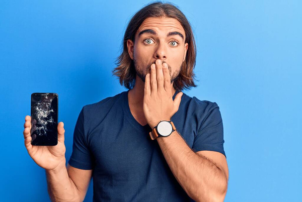 Mladý pohledný muž, držící rozbitý smartphone, ukazující popraskanou obrazovku zakrývající ústa rukou, šokovaný a vystrašený z omylu. překvapený výraz  - Fotografie, Obrázek