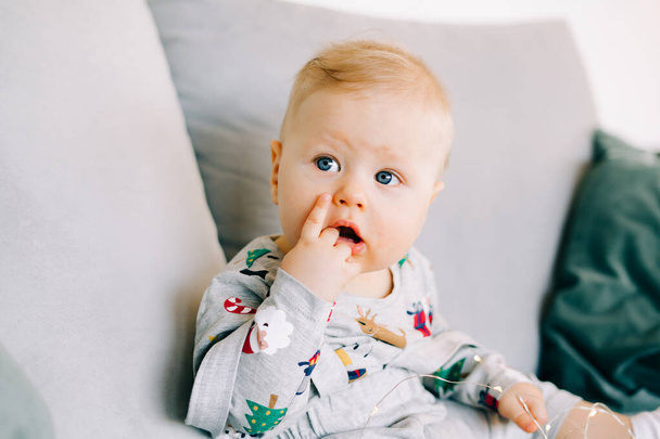 a cute little boy who is teething is sitting in a festive Christmas costume on a gray sofa and playing with a bright light garland.Christmas concept. - Valokuva, kuva
