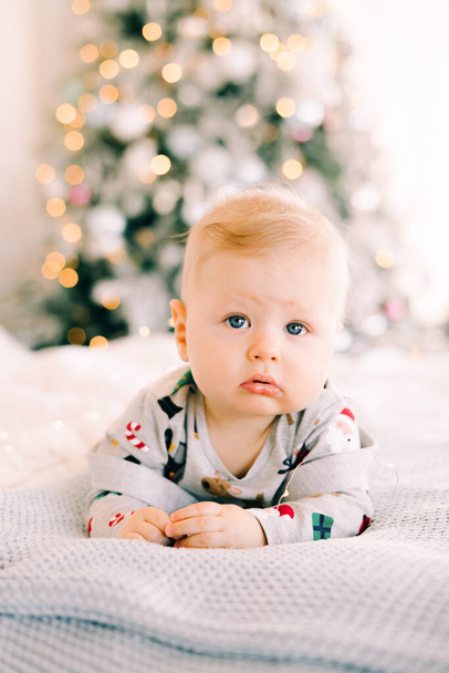 子供の最初のクリスマスだ。黄金の光のガーランドを持つ明るい装飾されたモミの木の背景にベッドの上のお祝いの衣装に横たわっています。クリスマスのコンセプト. - 写真・画像