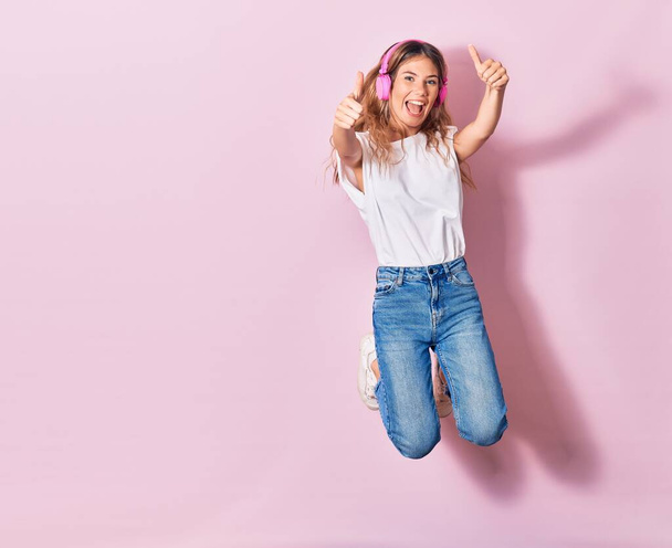 Young beautiful blonde woman smiling happy. Jumping with smile on face listening to music using pink headphones doing ok sign with thumbs up over isolated background - Φωτογραφία, εικόνα