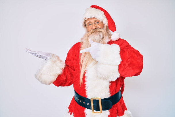 Vecchio uomo anziano con i capelli grigi e lunga barba indossando costume di Babbo Natale con bretelle stupito e sorridente alla fotocamera mentre si presenta con la mano e punta con il dito.  - Foto, immagini