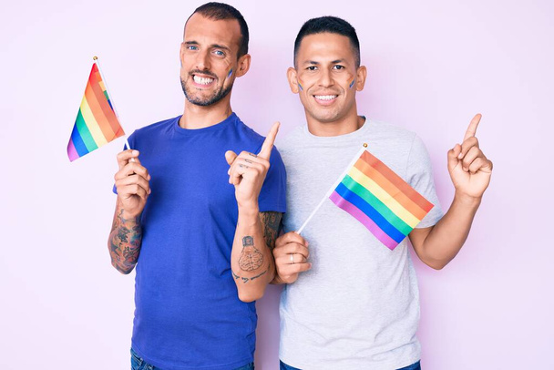 jong gay paar van twee mannen holding regenboog lgbtq vlaggen samen glimlachen gelukkig wijzend met hand en vinger naar de zijkant  - Foto, afbeelding