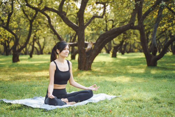 Young attractive woman meditating in lotus position sitting on the grass in the park. Yoga and harmony concept. - 写真・画像