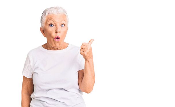 Senior beautiful woman with blue eyes and grey hair wearing casual white tshirt surprised pointing with hand finger to the side, open mouth amazed expression.  - Foto, imagen