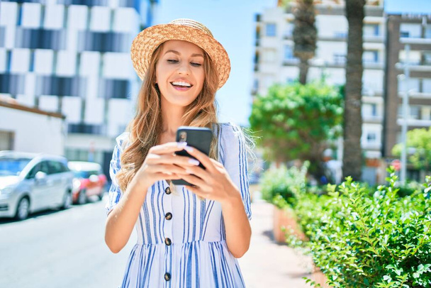 Νεαρή όμορφη ξανθιά γυναίκα σε διακοπές φορώντας καλοκαιρινό καπέλο χαμογελώντας ευτυχισμένη. Μόνιμη με χαμόγελο στο πρόσωπο χρησιμοποιώντας smartphone στο δρόμο της πόλης. - Φωτογραφία, εικόνα