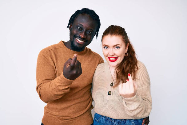 Couple Interracial portant des vêtements décontractés signe viennent ici geste avec la main invitant accueillant heureux et souriant  - Photo, image