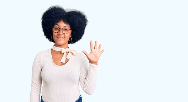 Молода афроамериканка в повсякденному одязі і окулярах показує і вказує вгору пальцями номер п'ять, посміхаючись впевнено і щасливо.  - Фото, зображення