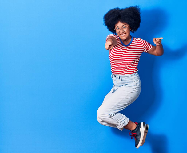 若い美しいアフリカ系アメリカ人女性のカジュアルな服や眼鏡を着て幸せな笑顔。孤立した青の背景の上に笑顔でジャンプ - 写真・画像