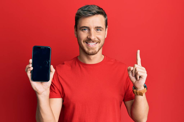 Bonito homem caucasiano segurando smartphone mostrando tela sorrindo com uma ideia ou pergunta apontando dedo com rosto feliz, número um  - Foto, Imagem