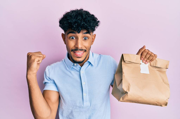 Young arab handsome man holding take away paper bag screaming proud, celebrating victory and success very excited with raised arm  - Фото, зображення