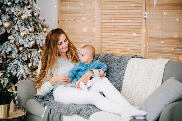 Baby girl, daughter with mom on sofa near Christmas tree. Happy New Year and Merry Christmas. Christmas decorated interior. The concept of family holiday. - Фото, изображение