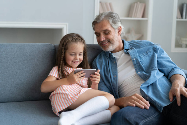 Cute girl help father show something on smartphone, smart little daughter and dad sit on couch hold mobile phone - 写真・画像