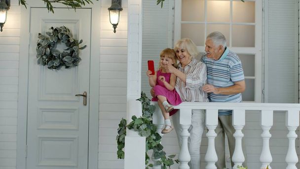 Pareja mayor sentada con su nieta en el porche de su casa. Uso del teléfono móvil para videollamadas en línea - Foto, Imagen