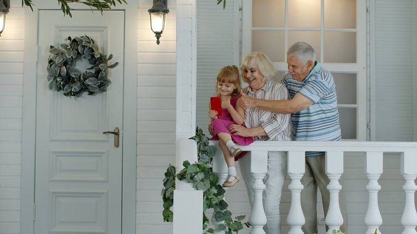 Oudere stel zit met kleindochter in de veranda thuis. Mobiele telefoon gebruiken voor online videogesprekken - Foto, afbeelding