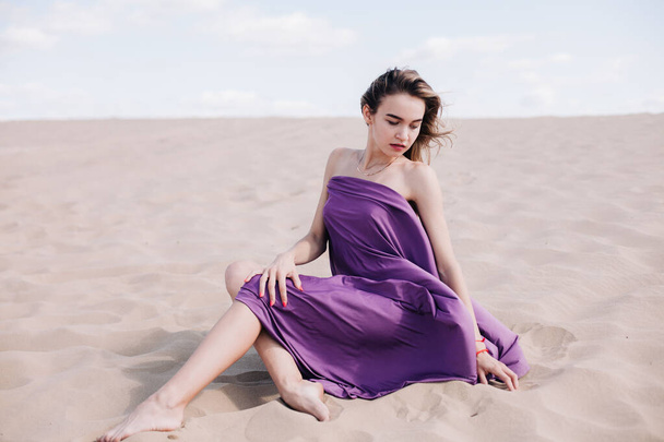 A young, slender girl in a beige dress with purple cloth in her hands posing in the desert in the wind - Fotó, kép