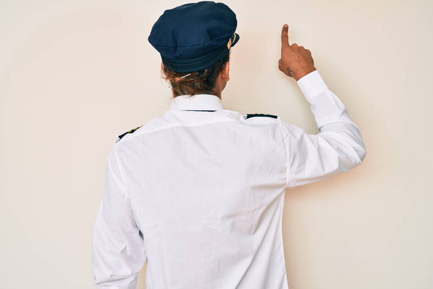 Young hispanic man wearing airplane pilot uniform posing backwards pointing ahead with finger hand  - Fotó, kép