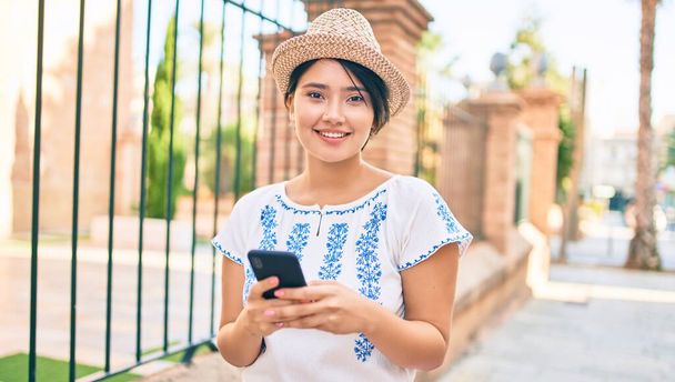 Νεαρή Λατίνα τουρίστρια κοπέλα στις διακοπές χαμογελώντας ευτυχισμένη χρησιμοποιώντας smartphone στην πόλη. - Φωτογραφία, εικόνα