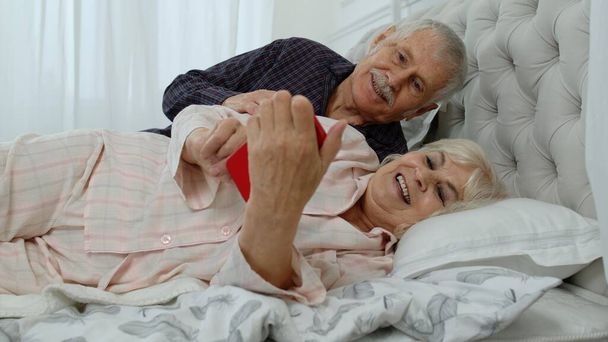 Пожилая пара в пижаме лежит на кровати и смотрит на мобильный телефон, смеясь и веселясь - Фото, изображение