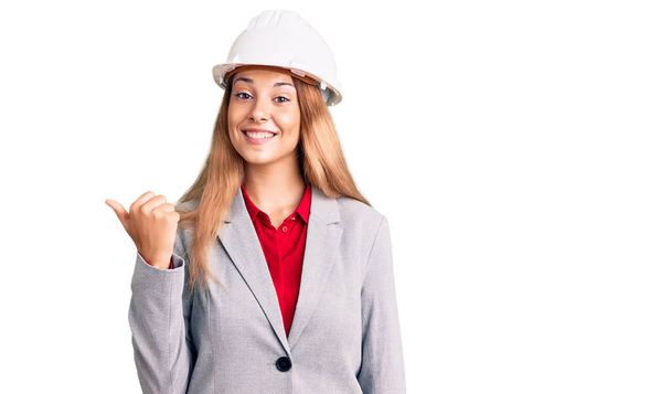 Beautiful young woman wearing architect hardhat smiling with happy face looking and pointing to the side with thumb up.  - Photo, Image