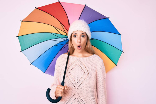 młoda brunetka kobieta pod kolorowy parasol przestraszony i zdumiony z otwartymi ustami dla zaskoczenia, niedowierzanie twarz  - Zdjęcie, obraz