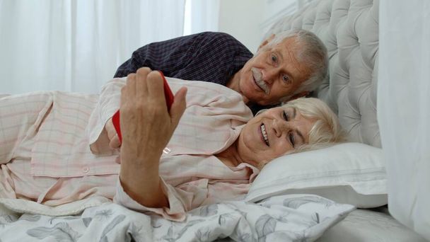 Starsza para w piżamach leżąca na łóżku, patrząca na telefon komórkowy, śmiejąca się i bawiąca - Zdjęcie, obraz