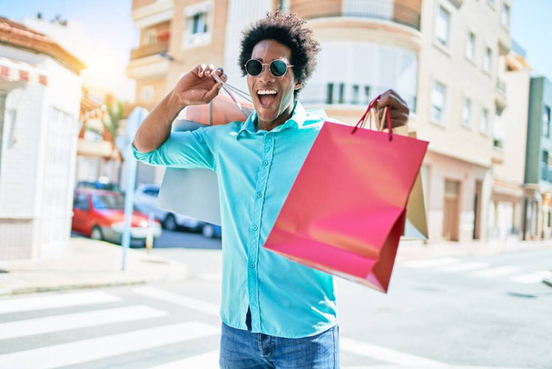 Young handsome african american man wearing sunglasses smiling happy. Standing with smile on face holding shopping bags at town street. - Foto, afbeelding