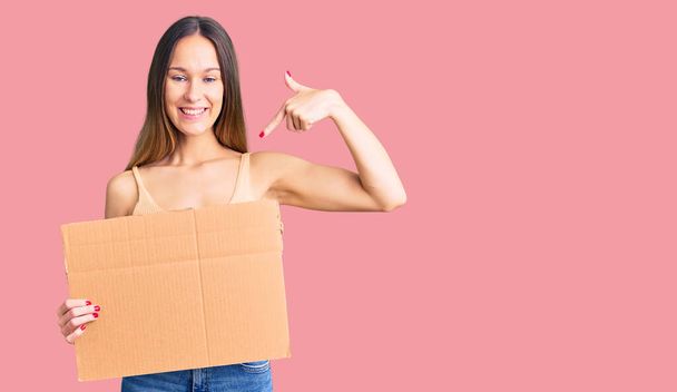 Belle jeune femme brune tenant une bannière en carton avec un espace vide pointant du doigt vers un sourire heureux et fier  - Photo, image