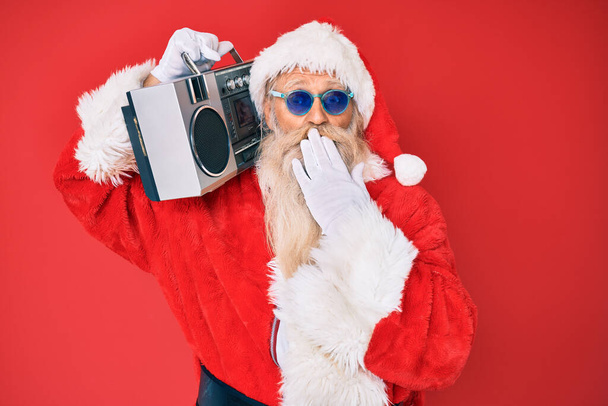 Old senior man wearing santa claus costume and boombox covering mouth with hand, shocked and afraid for mistake. surprised expression  - Fotoğraf, Görsel