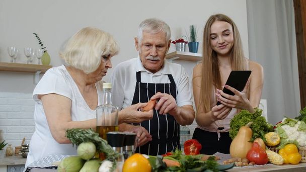 Ältere Großeltern schneiden Gemüse für Salat, hören Rezept von Mädchen mit Tablet - Foto, Bild