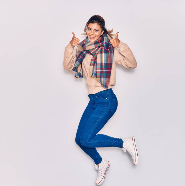 Young beautiful girl wearing winter clothes and scarf smiling happy. Jumping with smile on face doing ok sign with thumbs up over isolated white background - Photo, Image