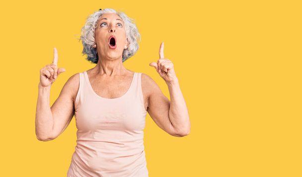 Senior grauhaarige Frau in lässiger Kleidung erstaunt und überrascht Blick nach oben und zeigt mit Fingern und erhobenen Armen.  - Foto, Bild