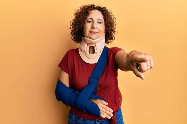 Beautiful middle age mature woman wearing cervical collar and arm on sling pointing with finger to the camera and to you, confident gesture looking serious  - Photo, Image