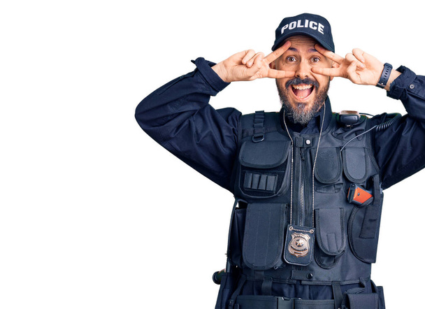 Jonge knappe man draagt politie-uniform doet vrede symbool met vingers over het gezicht, glimlachend vrolijk tonen overwinning  - Foto, afbeelding