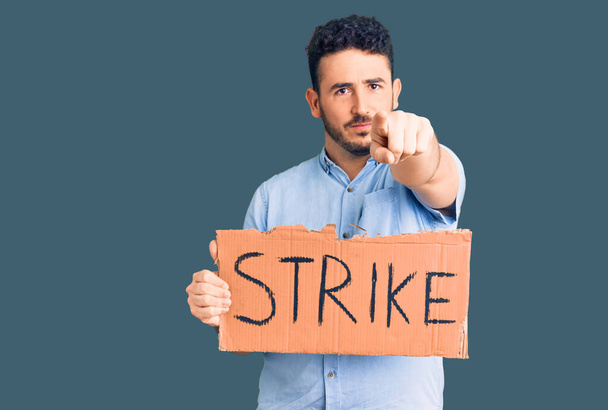 Young hispanic man holding strike banner cardboard pointing with finger to the camera and to you, confident gesture looking serious  - Photo, Image