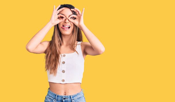 Young beautiful girl wearing casual sleeveless t shirt doing ok gesture like binoculars sticking tongue out, eyes looking through fingers. crazy expression.  - Photo, Image